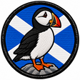Round 80mm embroidered patch showing a black and white puffin with an orange beak and feet standing on a rock against a Saltire, framed by a black border.