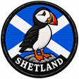 Round 80mm embroidered patch showing a puffin standing on a rock against a Saltire, with SHETLAND lettering along the bottom, framed by a black border.