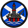 Round 80mm embroidered patch showing an offshore oil platform with a derrick and crane above waves, against a blue and white Saltire, framed by a black border.