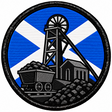 Round 80mm embroidered patch showing a coal mine pithead and winding wheel beside a coal wagon and heaps of coal, against a Saltire, framed by a black border.